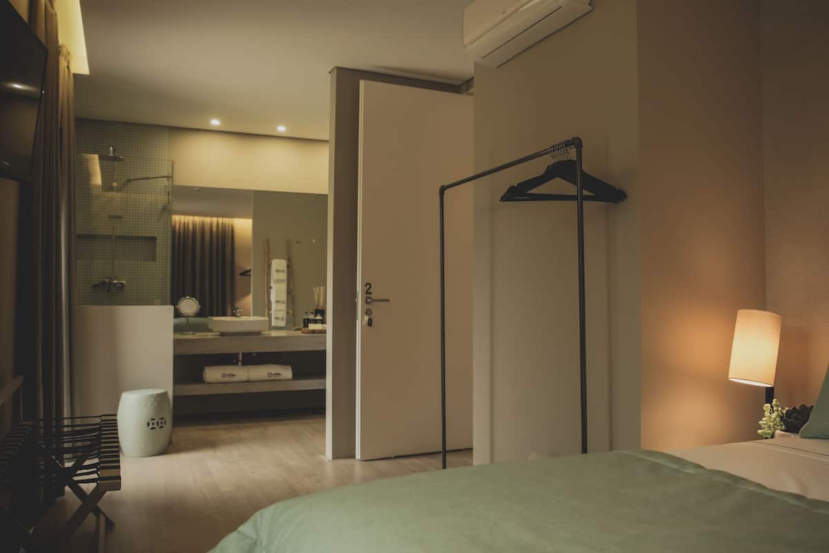 Quarto de hotel com cama, mesa de cabeceira, luminária, cabideiro, guarda-roupa, banheiro com pia e espelho, e vaso sanitário.