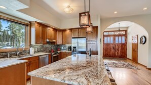 A spacious kitchen with wooden cabinets, a large marble island, and a view of the forest through the window. - Summit Sanctuary | Beautiful Views | Hot Tub! (Breckenridge)