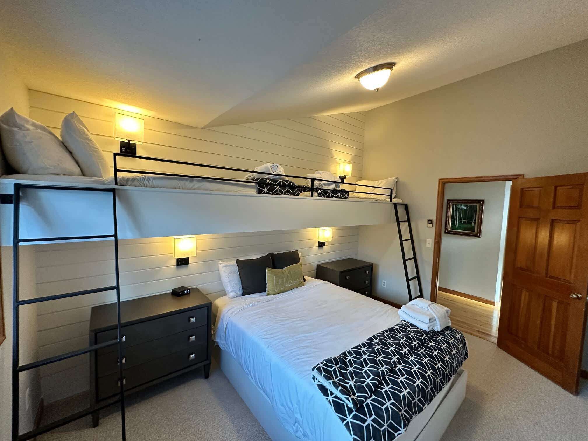 Une chambre avec un lit superposé, un bureau et une commode, une échelle et une porte ornée d’un tableau.