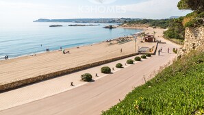 A coastal promenade with a stone pathway, beach, and a view of the sea and distant landforms.