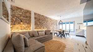 A modern living room with a stone wall, a sectional sofa, a dining area, and a kitchen. - Florida: Stylish Downtown Duplex with Sea Views (L'Escala)