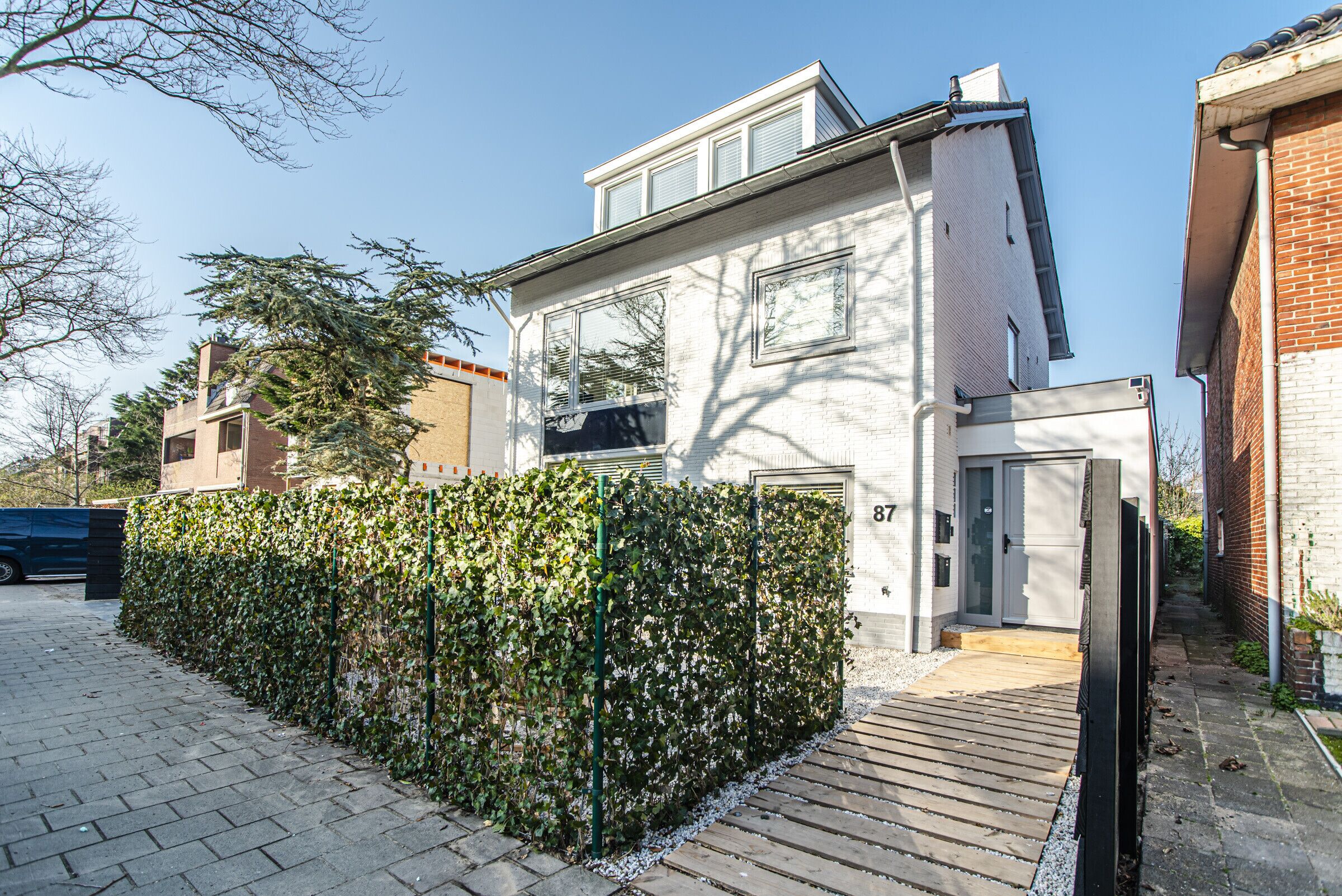 Una casa moderna de dos pisos con fachada blanca, terraza de madera y seto cuidado.