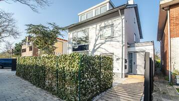 Una casa moderna de dos pisos con fachada blanca, terraza de madera y seto cuidado.