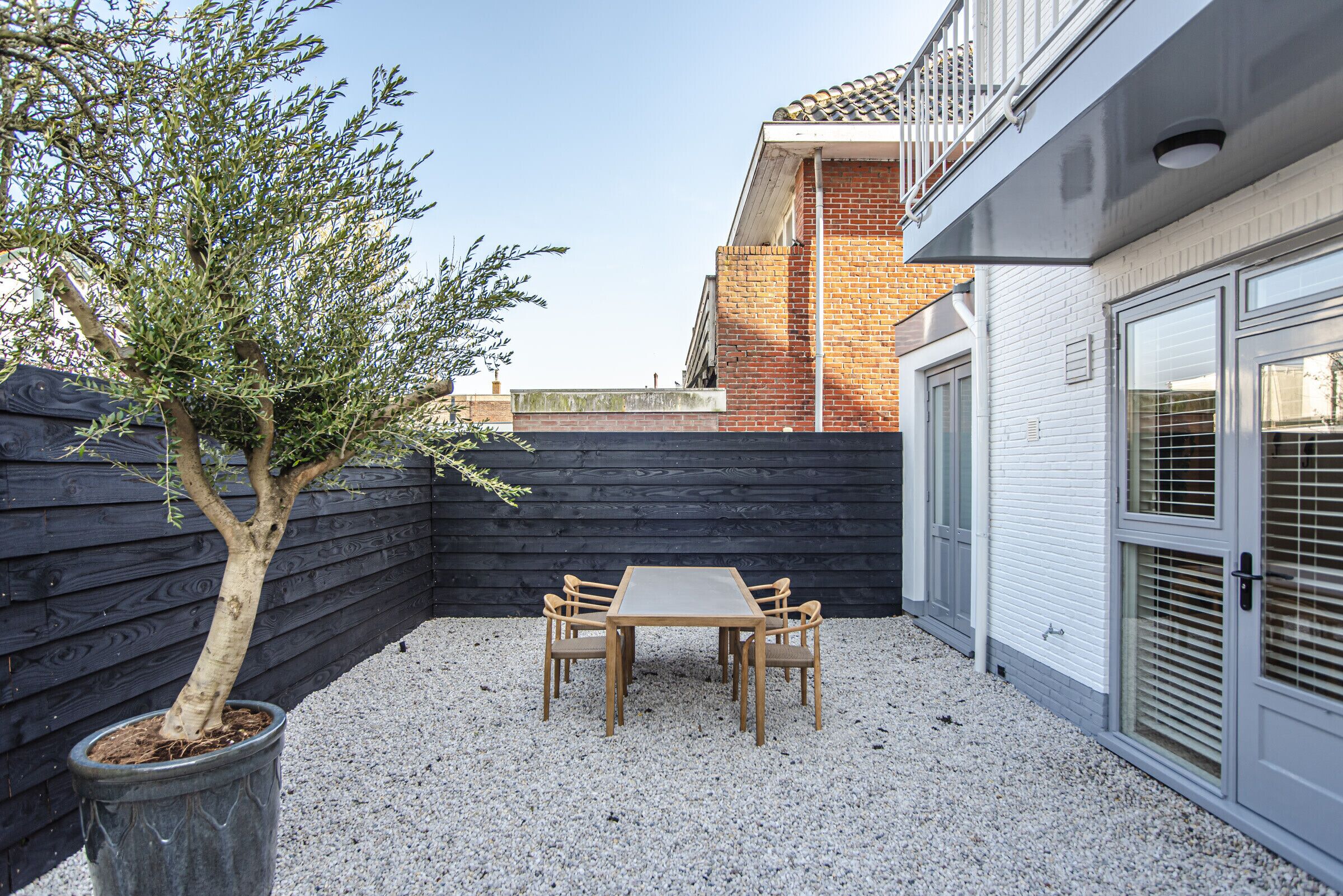 Eine Terrasse mit Tisch und Stühlen, ein Blumentopf mit einem Baum und ein Gebäude mit einer Ziegelwand und einer weißen Tür.