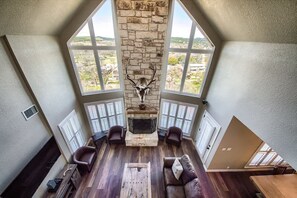 A loft-style room with a stone fireplace, leather sofas, a wooden coffee table, and large windows offering a view of the landscape. - CL 1181 Cypress Ridge (Canyon Lake)