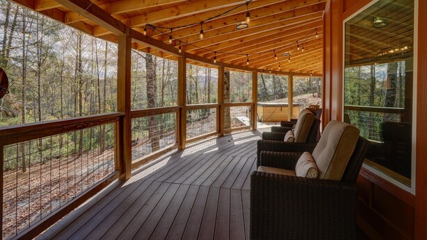 Une terrasse en bois, meublée de meubles en osier, avec de grandes fenêtres donnant sur une forêt.