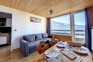 A modern room with a wooden ceiling, a grey sofa, a dining table set with plates, glasses, and cutlery, and a view of mountains through large windows.