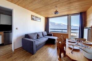 Una habitación acogedora con techo de madera, un sofá gris, una mesa de comedor con platos y cubiertos, y vistas a montañas a través de amplios ventanales.