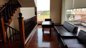 A spacious balcony with a wooden floor, a black sofa, and a small table. A wooden staircase with a railing leads to an upper level.