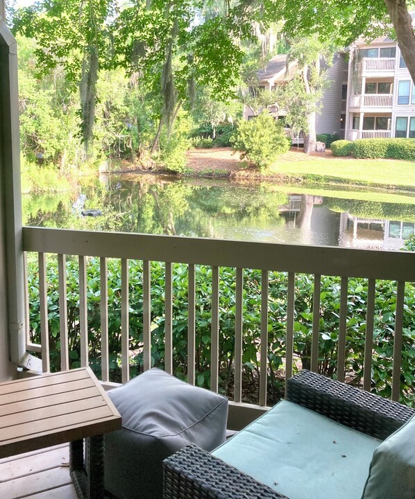 A balcony with a view of a pond, surrounded by greenery and a building in the background.