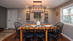 Uma sala de jantar com uma mesa de madeira, seis cadeiras, um candelabro, prateleiras para vinho e uma janela com vista.