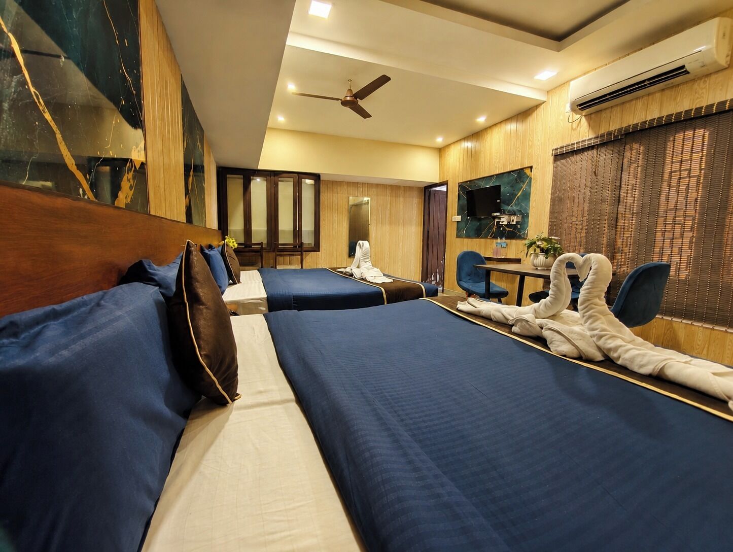 Quarto de hotel com duas camas, uma escrivaninha, uma cadeira, ventoinha no teto e uma janela grande com persianas.