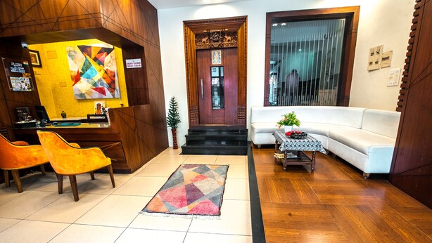 Um hall de hotel com uma mesa de recepção, duas cadeiras laranja, um sofá branco, um tapete estampado e piso de madeira.