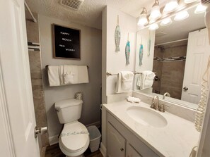 A bathroom with a toilet, sink, and a mirror with lights. There is a towel rack with towels and a sign above the toilet. - Sunrise Village 117 2 Bedroom Condo by RedAwning (Gulf Shores)