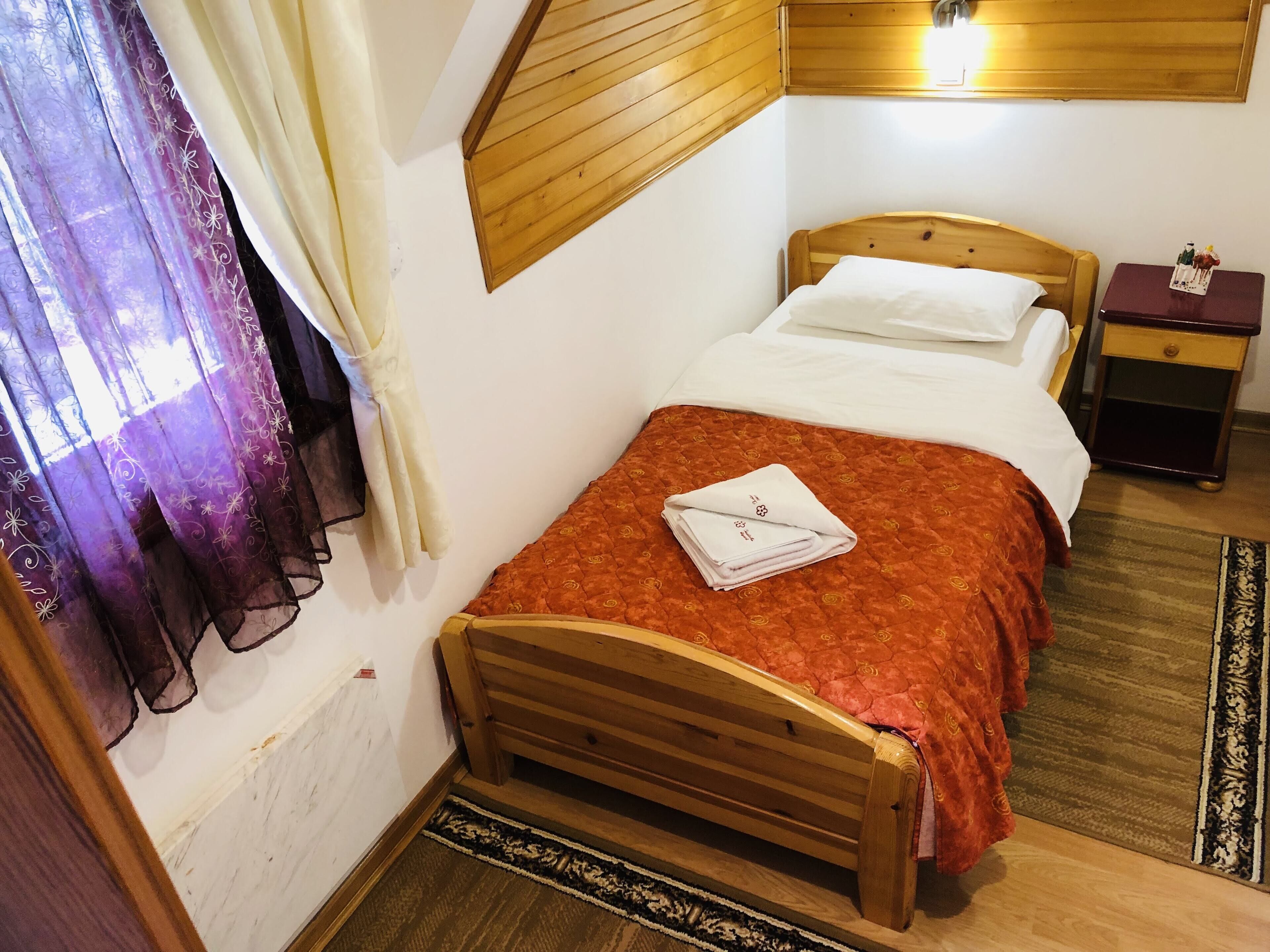 A single bed with a wooden headboard, a red patterned bedspread, and a folded white towel. There is a nightstand with a small figurine, a wooden headboard above the bed, and a window with purple curtains.