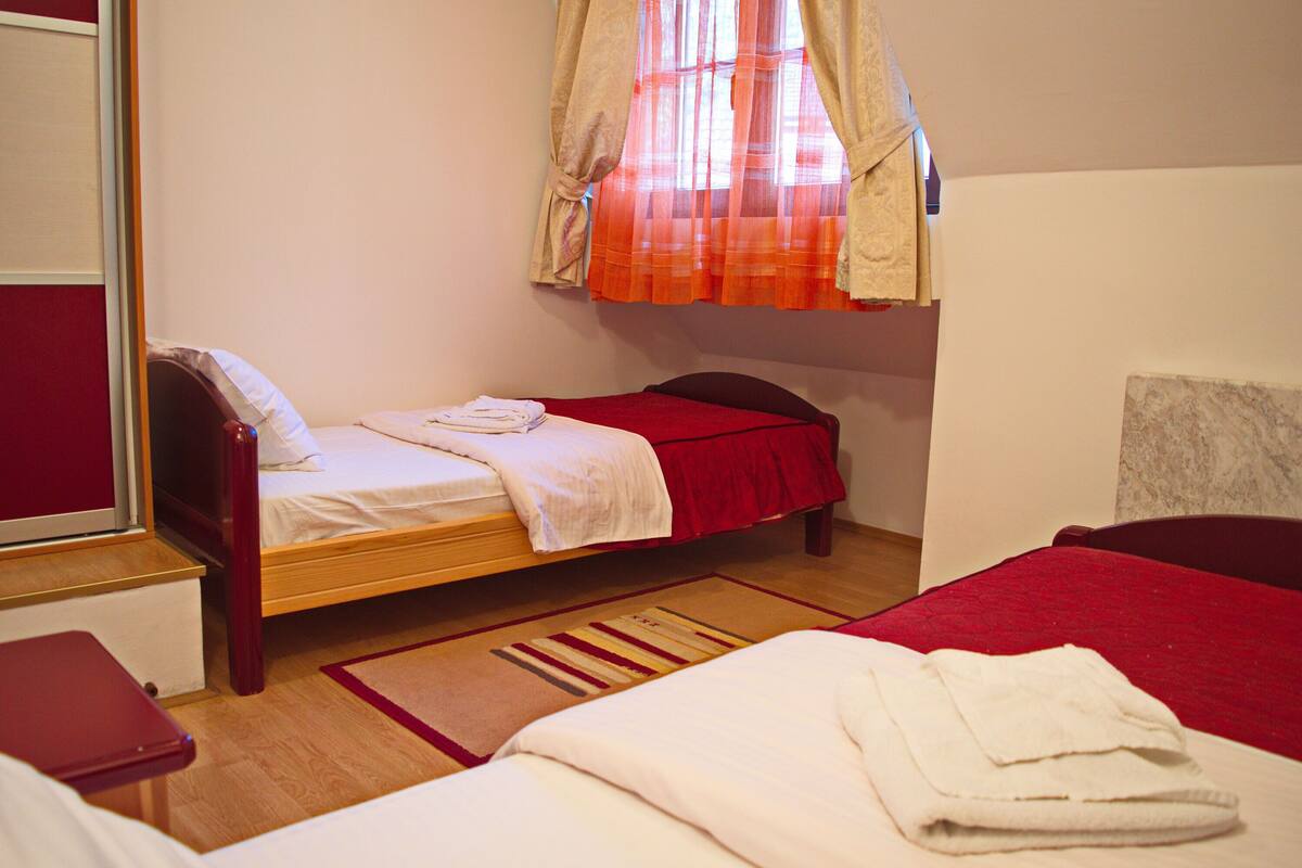 Une petite chambre avec un lit simple, un cadre de lit en bois, une couverture rouge et une fenêtre ornée de rideaux légers.