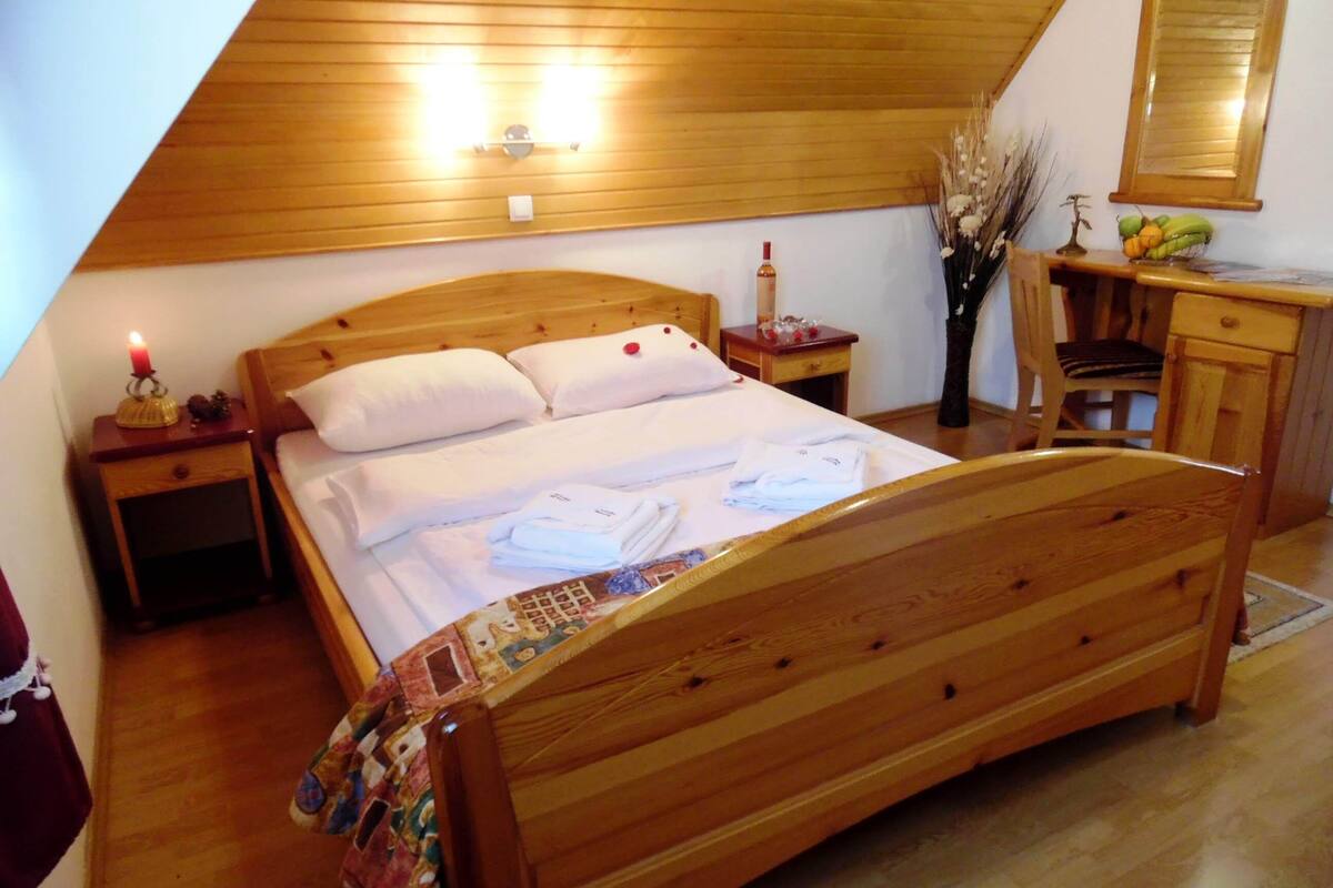 Un lit en bois, dans une pièce dont le plafond est en pente, avec une table de chevet sur laquelle se trouve une bougie, et un miroir au-dessus d’une commode.