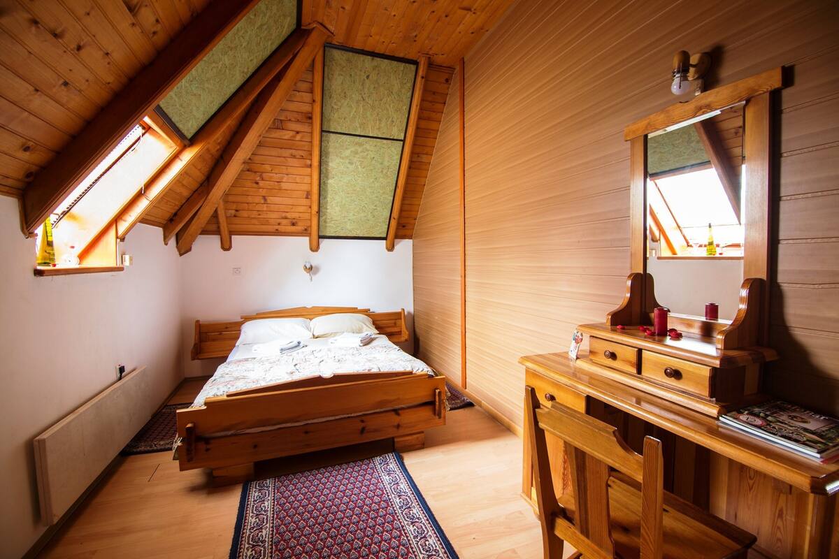 Une chambre avec un lit en bois, un bureau et un miroir. Il y a une lucarne au-dessus du lit et un radiateur au mur.