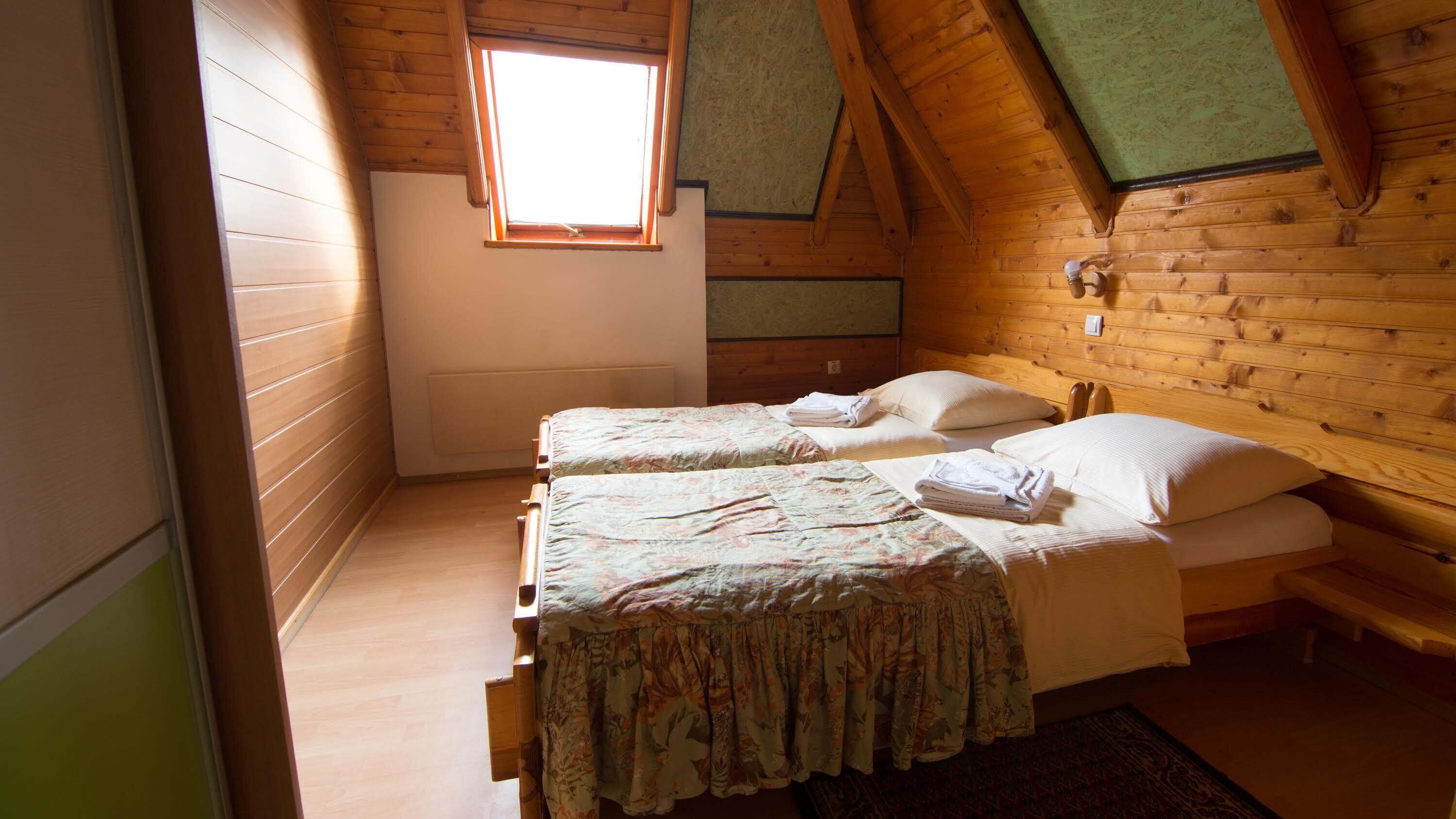 Una habitación pequeña y acogedora, con paredes de madera y techo inclinado. Hay dos camas con colchas estampadas con motivos florales y almohadas blancas. Una ventana pequeña deja entrar luz natural.