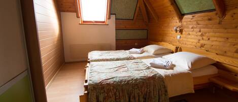 Una habitación pequeña y acogedora, con paredes de madera y techo inclinado. Hay dos camas con colchas estampadas con motivos florales y almohadas blancas. Una ventana pequeña deja entrar luz natural.
