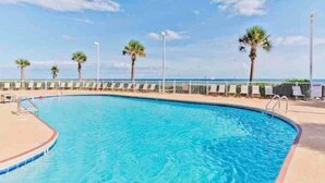 A swimming pool with a curved edge, surrounded by a white fence and palm trees. - Beachfront Condo with Spectacular Views and Heated Pool! (Orange Beach)