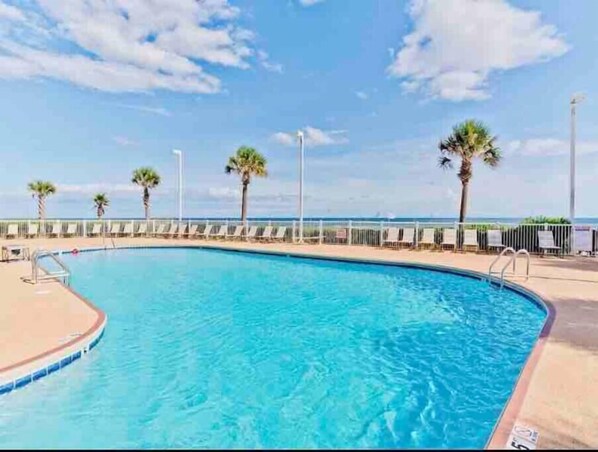 A swimming pool with a curved edge, surrounded by a white fence and palm trees. - Beachfront Condo with Spectacular Views and Heated Pool! (Orange Beach)
