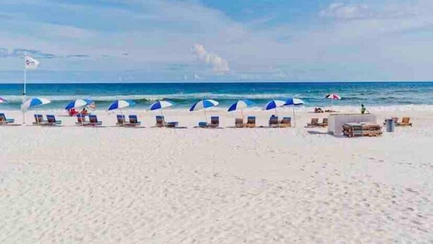 A beach with rows of sun loungers and umbrellas, a flag, and the ocean in the background. - Beachfront Condo with Spectacular Views and Heated Pool! (Orange Beach)
