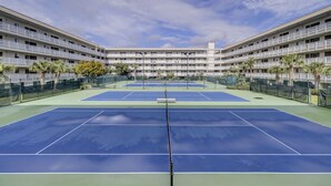 Tenniskenttä, jonka keskellä on verkko, ja jonka ympärillä on aita. Taustalla näkyy parvekkeilla varustettu monikerroksinen rakennus.