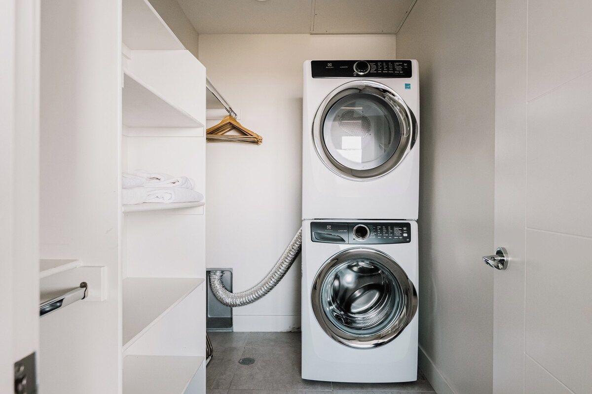 A compact laundry room with a front-loading washer and dryer, a hanging rack, and a white shelving unit.
