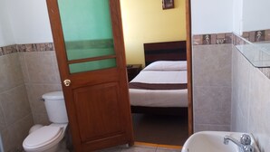 A bathroom with a toilet, a sink, and a bed visible through an open door. - EL MONARCA AREQUIPA (Arequipa)