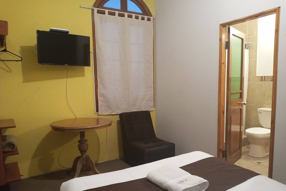 Habitación de hotel con cama, una silla, una mesita, televisor montado en la pared y un baño visible a través de una puerta abierta.