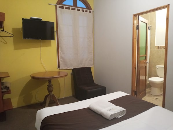 A hotel room with a bed, a chair, a small table, a TV mounted on the wall, and a bathroom visible through an open door.