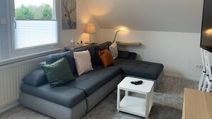Ein modernes Wohnzimmer mit einem grauen Ecksofa, einem weißen Beistelltisch, einer Lampe und einem Fernseher auf einem Schrank.