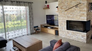 Un salon doté d’une cheminée en pierre, d’un téléviseur et offrant une vue sur un jardin grâce à de grandes fenêtres.
