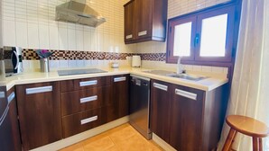 Een kleine keuken met donkere houten kastjes, een wit aanrecht, een inbouwvaatwasser en een raam met jaloezieën.