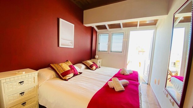 Een slaapkamer met een groot bed, witte nachtkastjes, een rode accentmuur en een raam met jaloezieën.