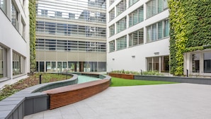 Un edificio moderno con un cortile che presenta una panca in legno curvo, fioriere e un piccolo elemento d'acqua.