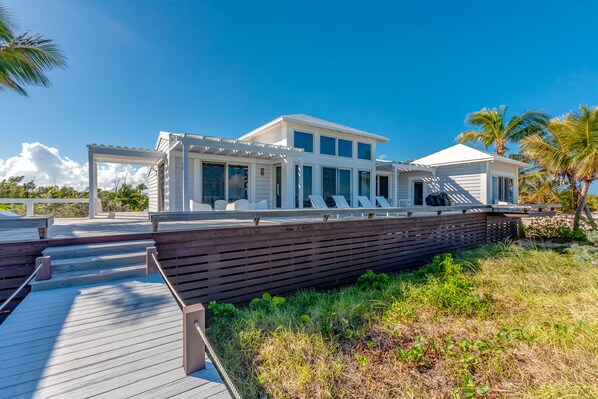 Una casa moderna frente a la playa con terraza de madera, ventanas grandes y un área de estar al aire libre techada.