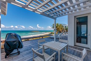 Un espacio al aire libre techado con piso de madera, sillas blancas y una mesa. Se ve una parrilla a la izquierda. La vista incluye una playa y el océano.