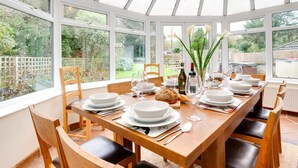 Une salle à manger aménagée dans une véranda, avec une table en bois dressée pour six personnes, entourée de chaises, et de grandes fenêtres donnant sur le jardin.