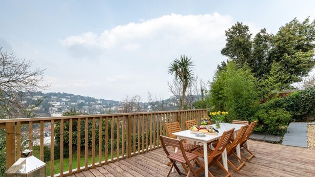 A wooden deck with a table set for dining, surrounded by chairs, overlooking a cityscape. - 5 Bed in Torquay (oc-bcvane) (Torquay)