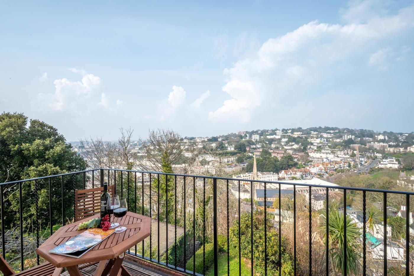 A balcony with a table, wine glasses, and a bottle, overlooking a cityscape with buildings and greenery.