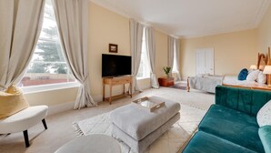 A spacious living room with a television, sofa, ottoman, and a view of a building outside. - 5 Bed in Torquay (oc-bcvane) (Torquay)
