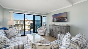 Sala de estar con un sofá, una mesa de centro, una silla y un mueble. Se ve una ventana grande con vistas a edificios y un balcón.