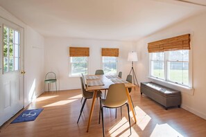 A room with a wooden floor, a dining table with chairs, a bench, and two windows with blinds.