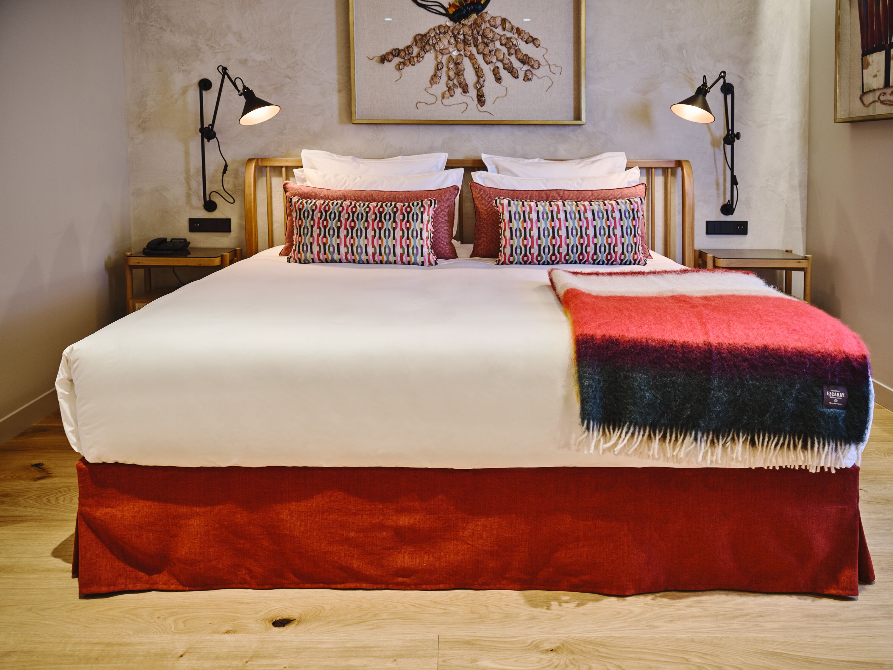Una cama bien hecha con una colcha blanca y roja, dos almohadas estampadas y una manta roja. Hay dos mesitas de noche con lámparas, una obra de arte enmarcada sobre la cama y un suelo de madera.