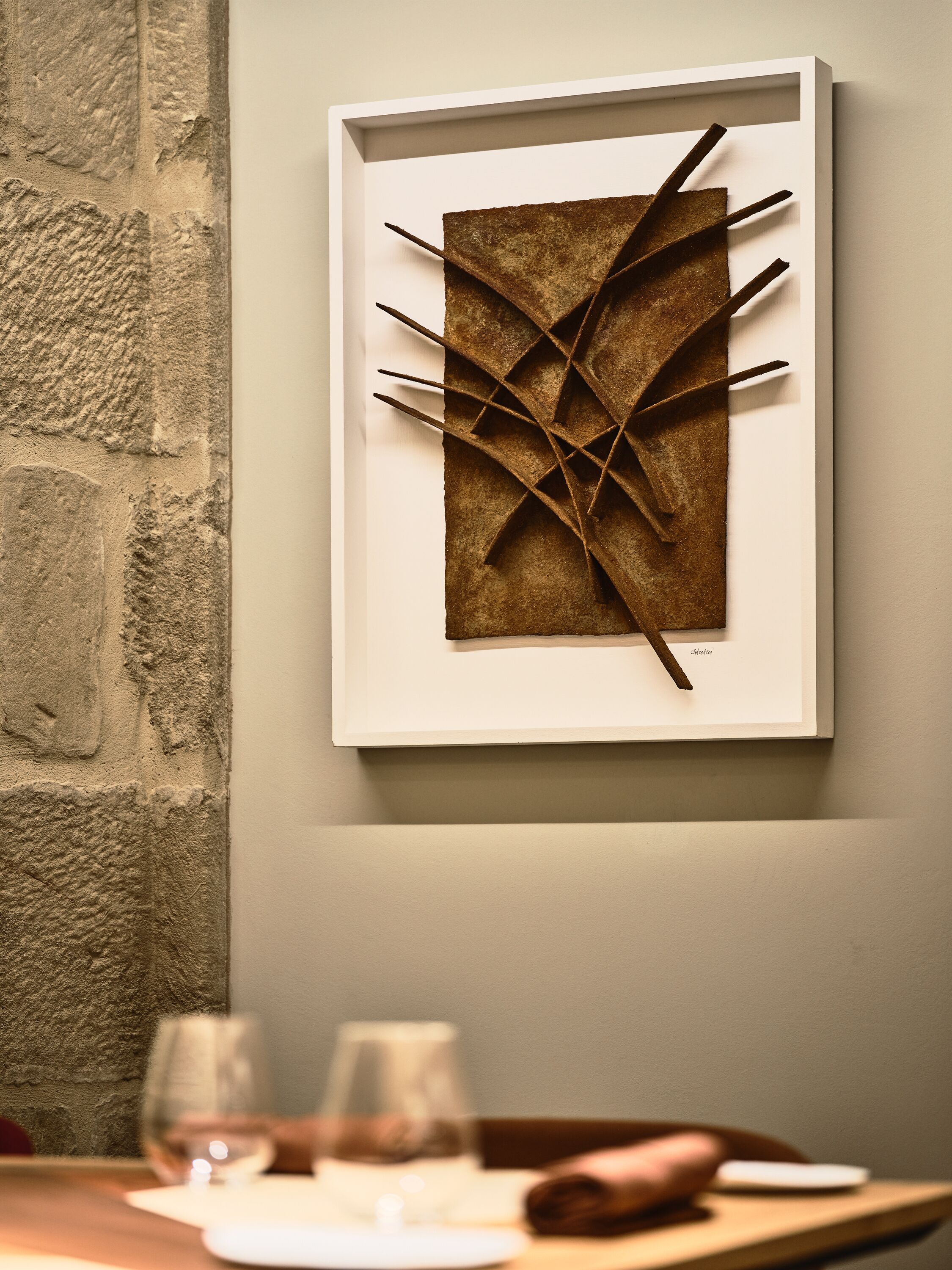 A framed abstract artwork on a wall, a textured stone wall, and a table with glasses and a napkin.