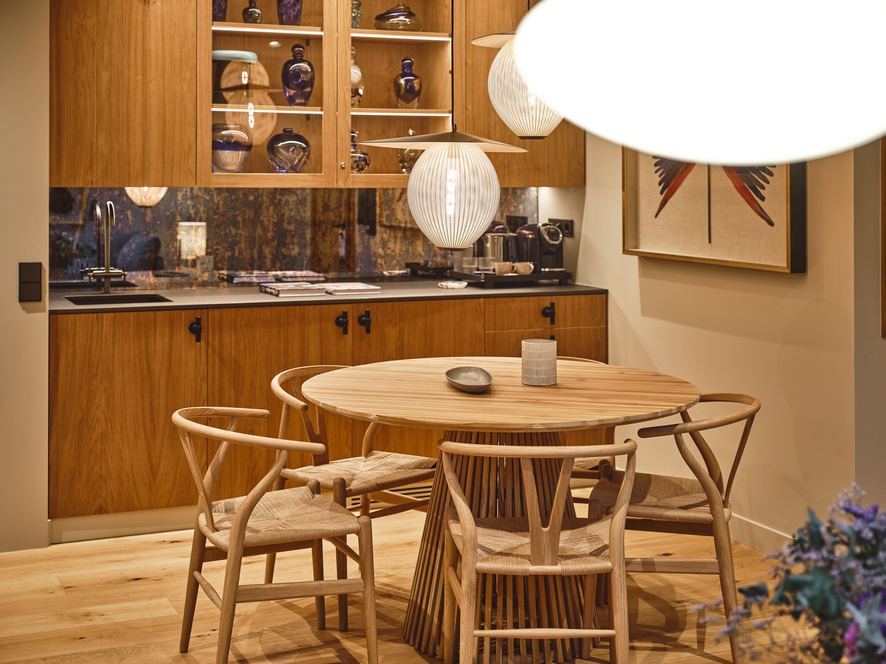 Un comedor de madera con una mesa redonda y cuatro sillas, un aparador con puertas de cristal que exhibe varios objetos, y un colgador de luz.