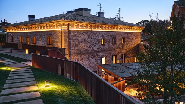 Un edificio de piedra con techo de tejas, terraza de madera y escalera que lleva a un espacio exterior cubierto.