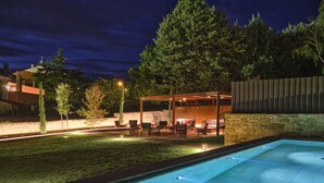 Zona de piscina exterior de noche con un muro de piedra, un área de estar cubierta y un césped bien iluminado.